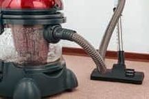 Industrial powered steam cleaner displayed on a beige carpet in a living room.  The steam cleaner has a red top and the tank is clear displaying the water inside.
